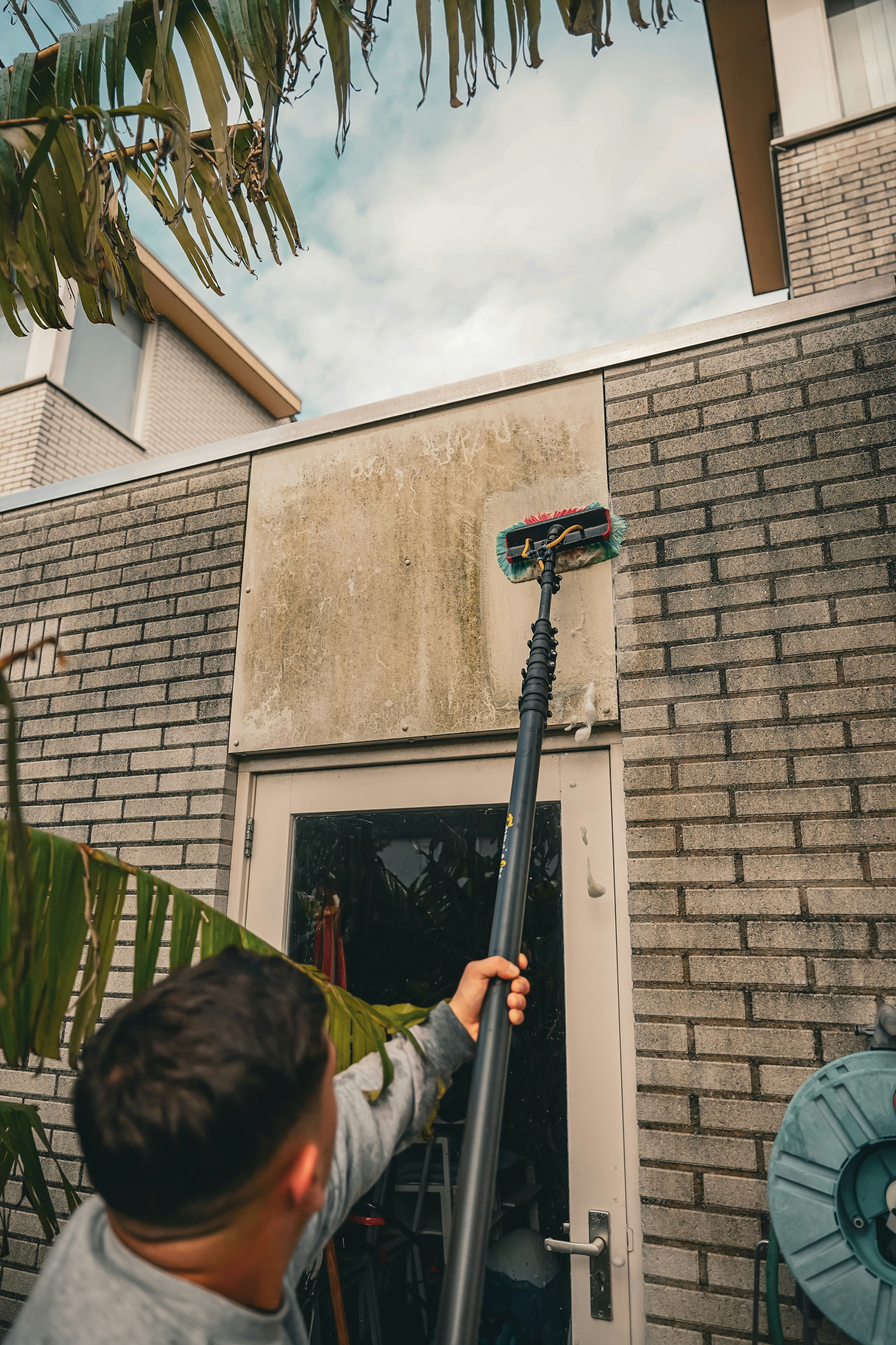 A person cleaning a grimy exterior wall with a long handled brush