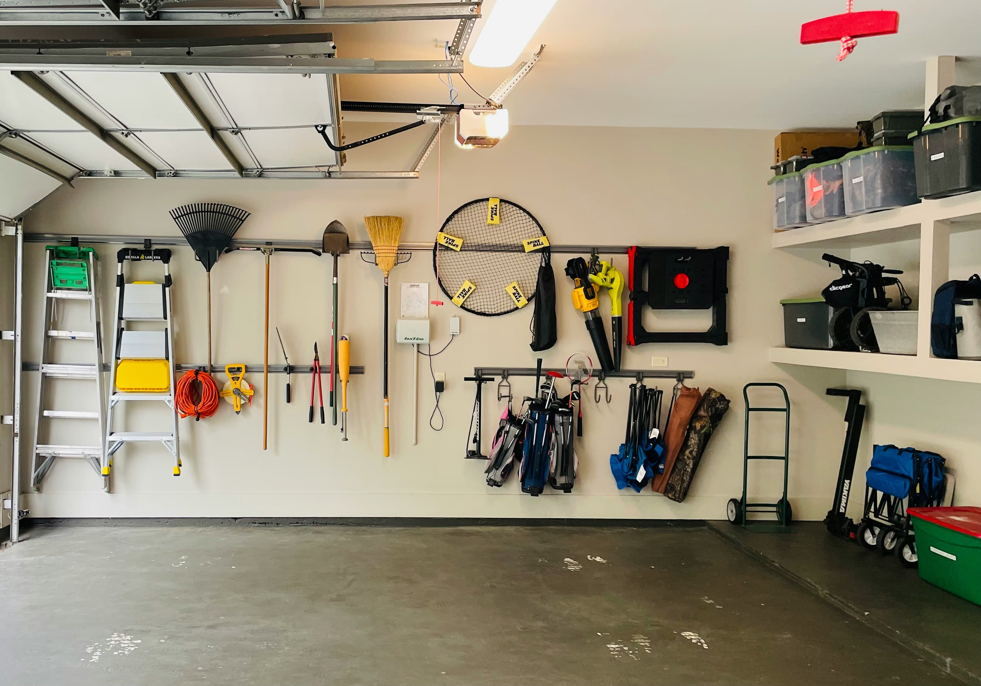 A tidy garage wall neatly hung with a ladder, rake, leaf blower, golf clubs, folding chairs, and a hand truck